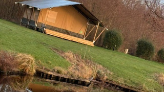 Una tenda safari si trova su un prato verde vicino a uno stagno, con alberi sullo sfondo e cielo sereno.