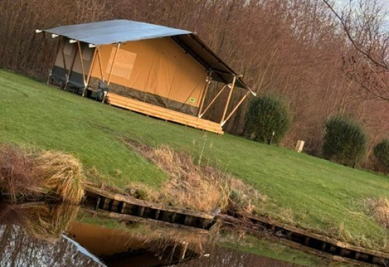 Una tenda safari si trova su un prato verde vicino a uno stagno, con alberi sullo sfondo e cielo sereno.