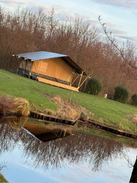 Een safaritent staat op het gras aan een vijver, omgeven door bomen en natuur op een heldere dag.