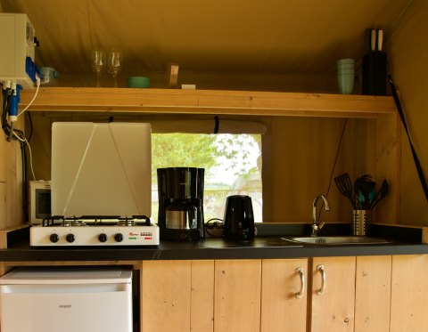 Kitchen area inside a safari tent with stove, fridge, coffee maker, sink, and various utensils.