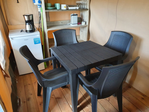 Interior de una tienda safari con mesa y sillas negras de plástico, frigorífico, cafetera y estantería.
