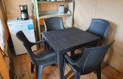 Interior de una tienda safari con mesa y sillas negras de plástico, frigorífico, cafetera y estantería.