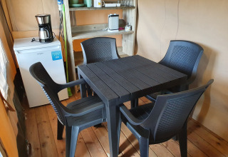 Interior de una tienda safari con mesa y sillas negras de plástico, frigorífico, cafetera y estantería.