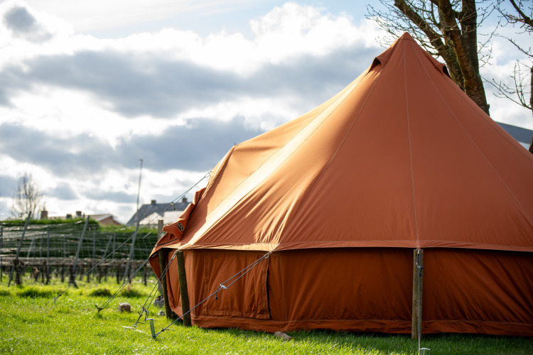 Gran tienda de campaña marrón para glamping en un campo verde con casas y cielo nublado al fondo.