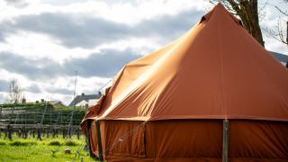 Gran tienda de campaña marrón para glamping en un campo verde con casas y cielo nublado al fondo.