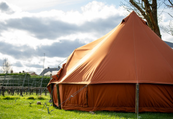 Grande tenda marrone da glamping su prato verde, con case e cielo nuvoloso sullo sfondo durante il giorno.