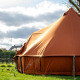 Grote bruine glampingtent op een groene weide met bewolkte lucht en huizen op de achtergrond overdag.