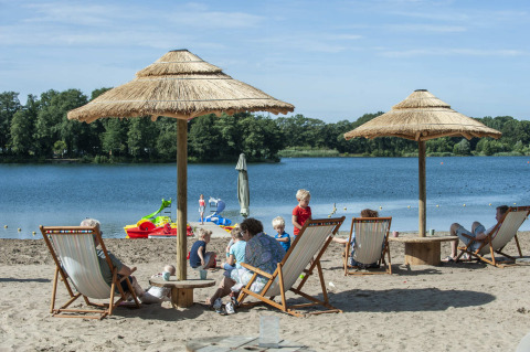 Camping Sevink Molen in Gelderland, Niederlande, mit Menschen, die am Seeufer unter Sonnenschirmen entspannen.