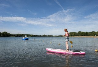 Girl stands on pink paddleboard at Camping Sevink Molen, Gelderland, Netherlands, under a clear sky.