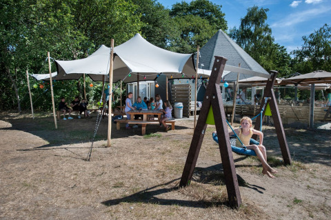 Børn leger på en gynge ved en legeplads med familier på picnic under telte, Camping Sevink Molen, Gelderland.