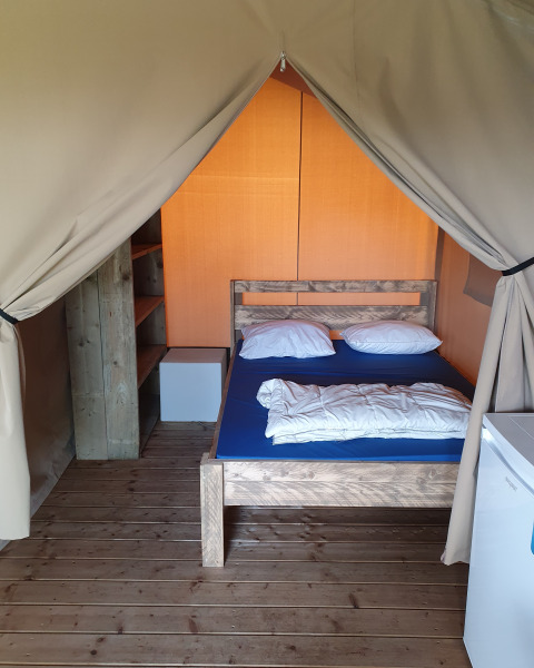 Vista interior de una tienda safari en Camping la Roche, Bélgica, con cama, estantería y suelo de madera.