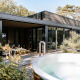 Moderne lodge med stor terrasse og udendørs boblebad, omgivet af frodig natur og grønne planter.