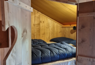 Acogedora habitación de madera con cama y edredón azul en tienda Safari con sanitario en Camping le Roc, Francia.