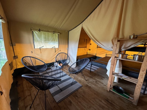 Vista interior de una tienda safari en Camping De La Vallée, Bélgica, con sillas, mesa y estantería de cocina.