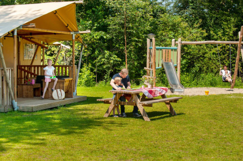 Familie genießt eine Pause vor einem Safarizelt auf Camping Sevink Molen, Kinder spielen auf dem Spielplatz.
