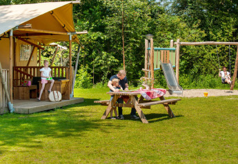 Famiglia rilassata davanti a una tenda safari al Camping Sevink Molen, bambini che giocano sul parco giochi.