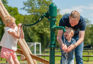 A man helps two children pump water from a green hand pump at Camping Sevink Molen in Gelderland, Netherlands.