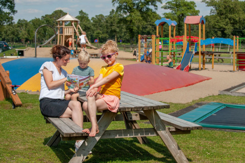 Madre y dos niños en banco junto a parque infantil, Camping Sevink Molen, Gelderland, Países Bajos.