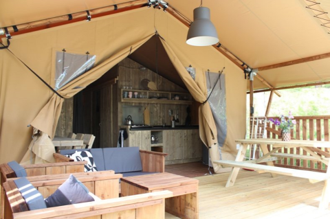 Interno tenda safari con arredi in legno, tavolo da picnic, ingresso aperto e cucina in fondo.