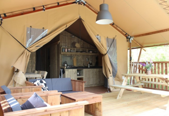 Intérieur de tente safari avec mobilier en bois, table de pique-nique, entrée ouverte et cuisine visible.