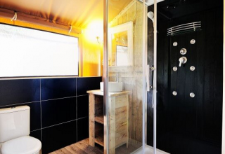 Baño moderno en tienda safari en Lac du Causse, Francia, con ducha, inodoro, lavabo y suelo de madera.