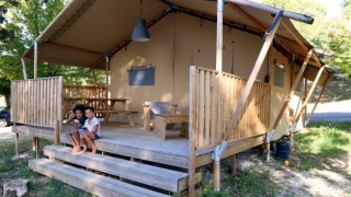 Twee kinderen zitten op de trap van een safaritent met houten veranda in een groene omgeving.