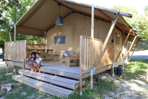 Due bambini seduti sui gradini di una tenda safari con veranda in legno, immersa nel verde.