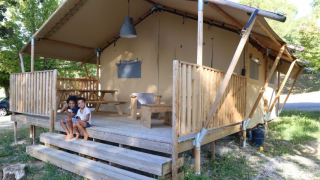 Due bambini seduti sui gradini di una tenda safari con veranda in legno, immersa nel verde.