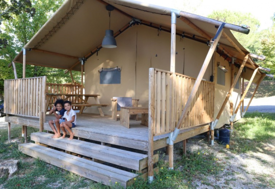 Two children sit on the steps of a safari tent with a wooden porch, surrounded by greenery outdoors.