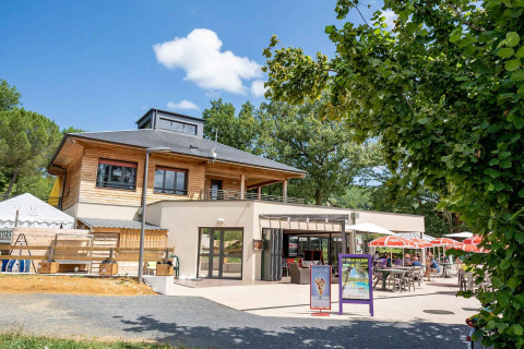 Udendørs café ved Lac du Causse feriepark med solskærme og gæster, omgivet af grønne træer i Frankrig.