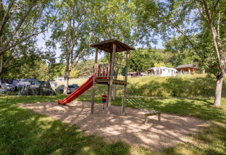 Parque infantil con tobogán y estructura de escalar en Lac du Causse, rodeado de árboles y casas móviles.