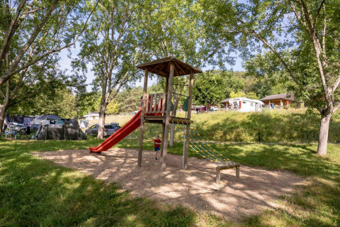 Parque infantil con tobogán y estructura de escalar en Lac du Causse, rodeado de árboles y casas móviles.