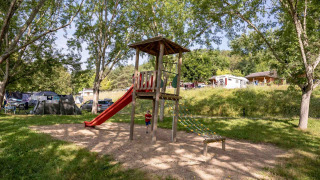 Parque infantil con tobogán y estructura de escalar en Lac du Causse, rodeado de árboles y casas móviles.