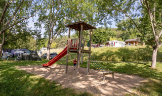 Parque infantil con tobogán y estructura de escalar en Lac du Causse, rodeado de árboles y casas móviles.