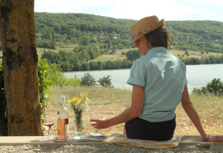 Vrouw met hoed geniet van wijn en uitzicht over meer en heuvels aan het Lac du Causse, Nouvelle-Aquitaine, Frankrijk.