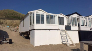 Strandhus 'Dishoek 345/346' med hvide vægge og store vinduer, placeret på sand ved klitter under blå himmel.