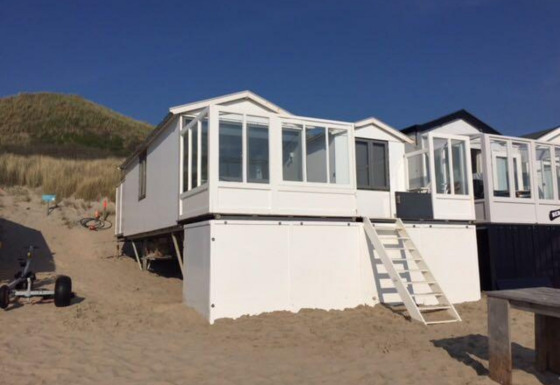 Maison de plage 'Dishoek 345/346' blanche avec véranda vitrée, posée sur le sable au pied d’une dune herbeuse.