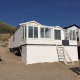 Strandhus 'Dishoek 345/346' med hvide vægge og store vinduer, placeret på sand ved klitter under blå himmel.
