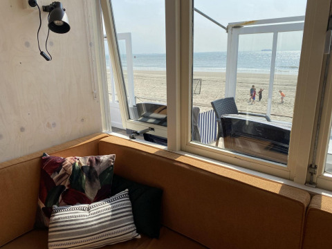 Vista desde una cabaña con sofá marrón y cojines hacia la playa y el mar en Dishoek, Zeeland, Países Bajos.