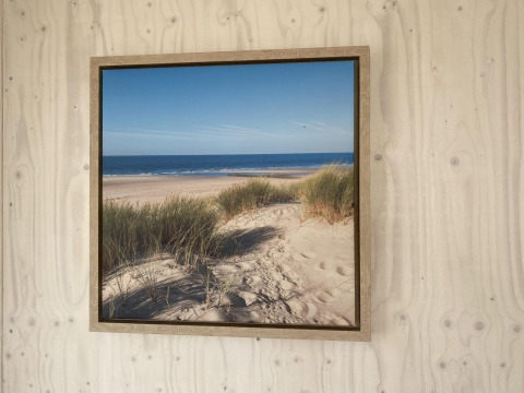 Cuadro enmarcado de playa de arena y dunas junto al mar, colgado en pared de madera en Beach house Dishoek.