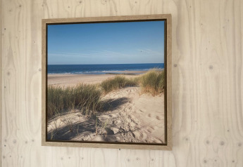 Cuadro enmarcado de playa de arena y dunas junto al mar, colgado en pared de madera en Beach house Dishoek.