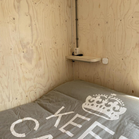 Cama pequeña con cobertor gris junto a una pared y repisa de madera en una cabaña en Dishoek, Países Bajos.