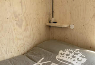 Cama pequeña con cobertor gris junto a una pared y repisa de madera en una cabaña en Dishoek, Países Bajos.