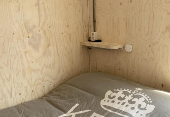 Cama pequeña con cobertor gris junto a una pared y repisa de madera en una cabaña en Dishoek, Países Bajos.