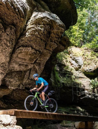 Cyclist crossing a wooden bridge beneath large rocky cliffs at Camping du Rivage in Diekirch, Luxembourg.
