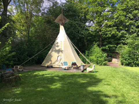 Una foto de la Tipi tent en un área verde, rodeada de árboles, con sillas y una mesa al frente.