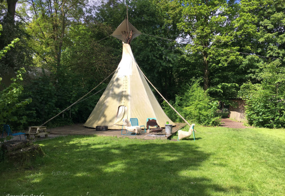 Una foto de la Tipi tent en un área verde, rodeada de árboles, con sillas y una mesa al frente.