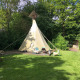 Et billede af en Tipi tent i en grøn have omgivet af træer, med stole og et bord foran teltet.