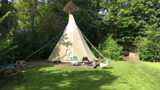 Una foto de la Tipi tent en un área verde, rodeada de árboles, con sillas y una mesa al frente.