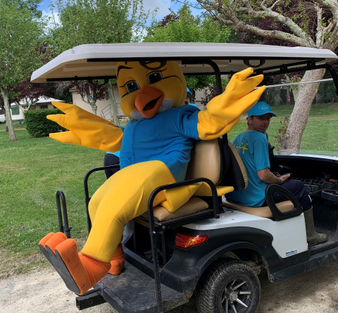 Mascota disfrazada de pájaro amarillo en camiseta azul en carrito de golf en Camping Paradis Aubeterre, Francia.
